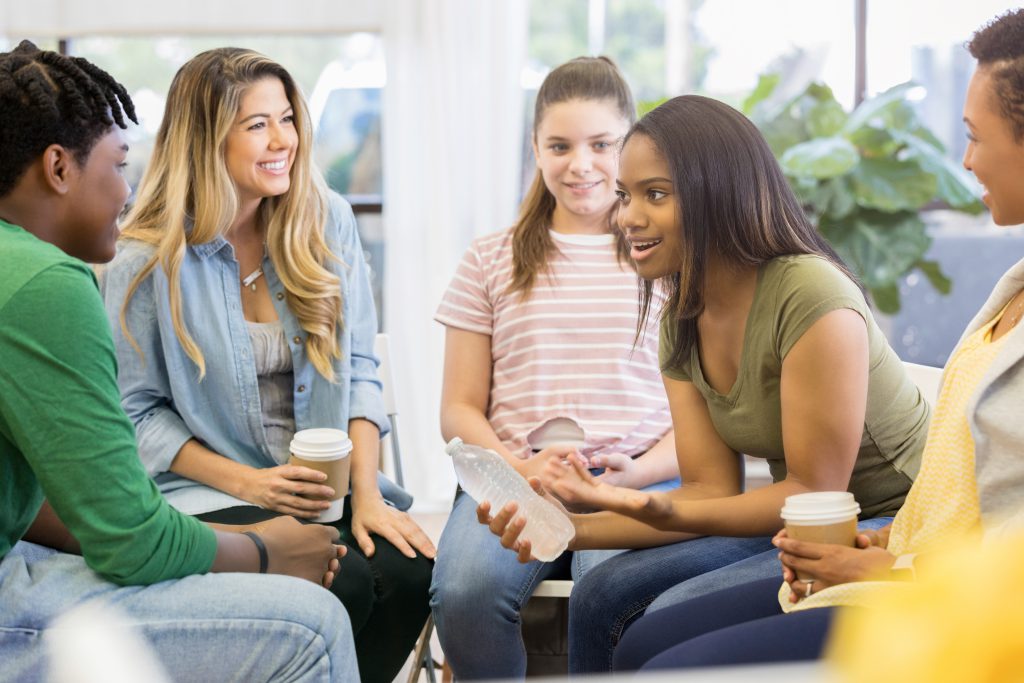 A young woman sits in a circle with her counseling support group.  She holds a water bottle as she leans forward and shares a humorous story.