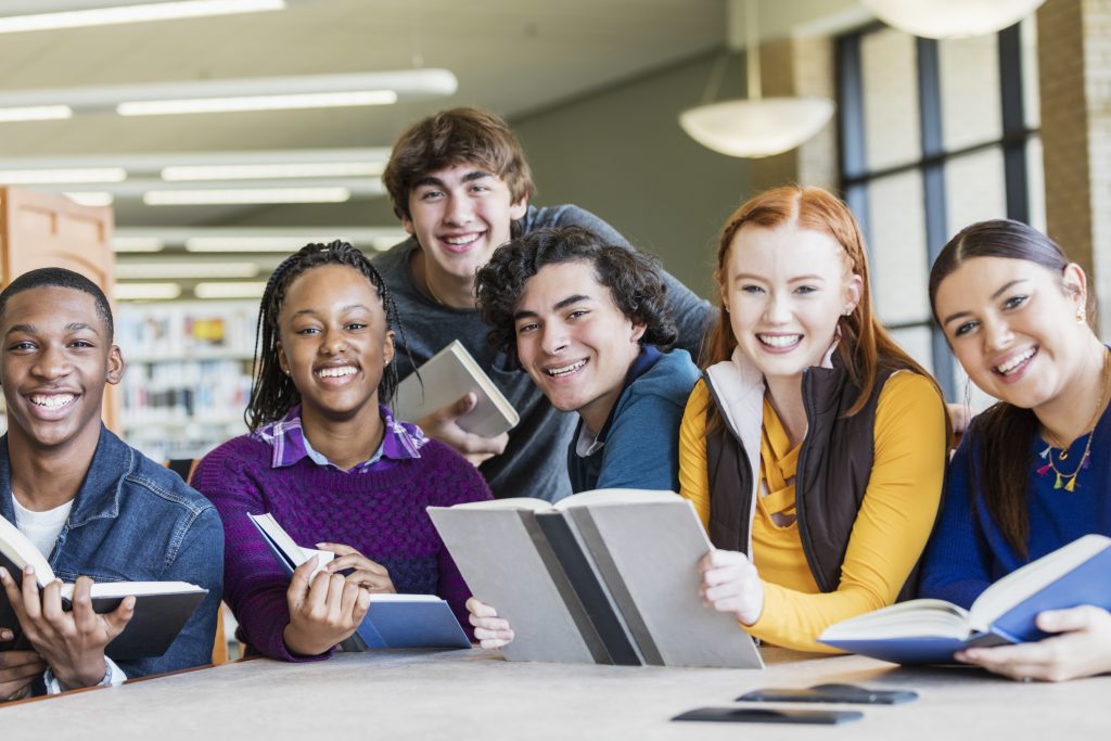 A multi-ethnic group of six high school students, 15 to 17 years old, studying together in the library. They are sitting at a table smiling at the camera.