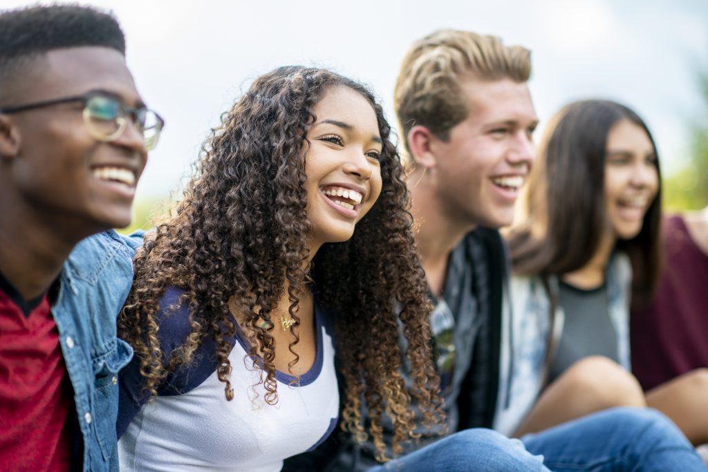 A girl of African American descent smiles with laughter while sitting in a row of teens with the arms around each other's shoulders. They are laughing too.