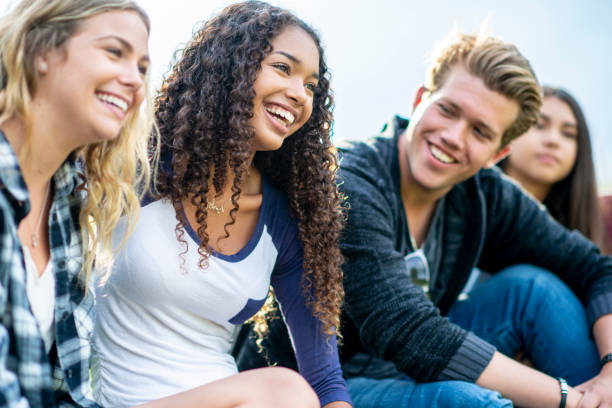 Two teen girls smile and laugh while they look into the distance, and a teenaged boy sits next to them and smiles while looking at them. They are sitting outside.