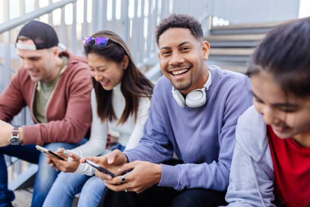 Young group of multiracial teenage people using mobile phone devices sitting outdoors. Young cheerful Latin American man holding smartphone and smiling at camera while relaxing with friends. Technology lifestyle and people concept